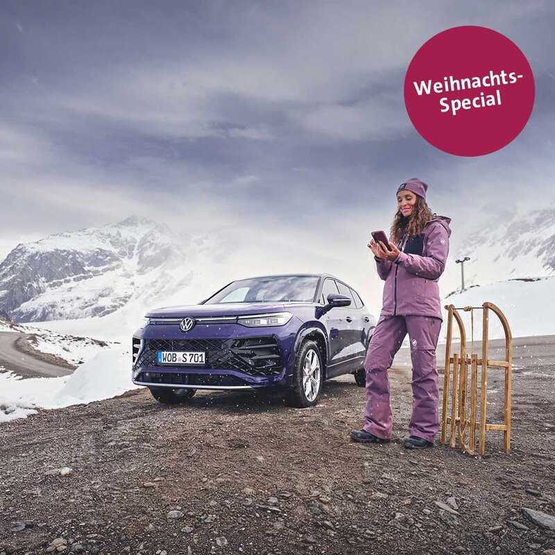 Frau in Winterkleidung steht neben einem VW Tayron in verschneiter Berglandschaft und hält ein Smartphone in der Hand. Daneben steht ein Holzschlitten.